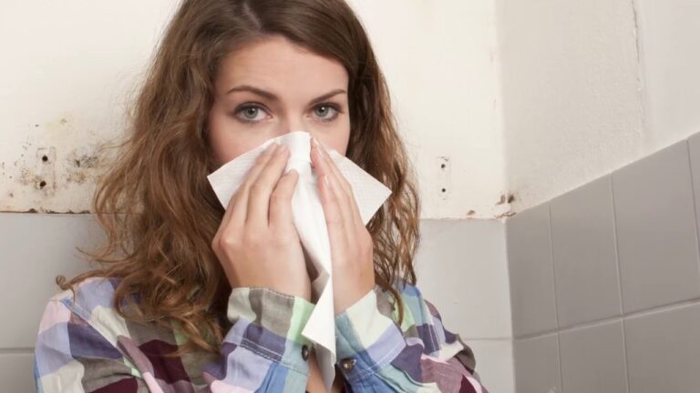 a sick woman covering her nose with tissue paper and mold can be seen in the background on the wall.